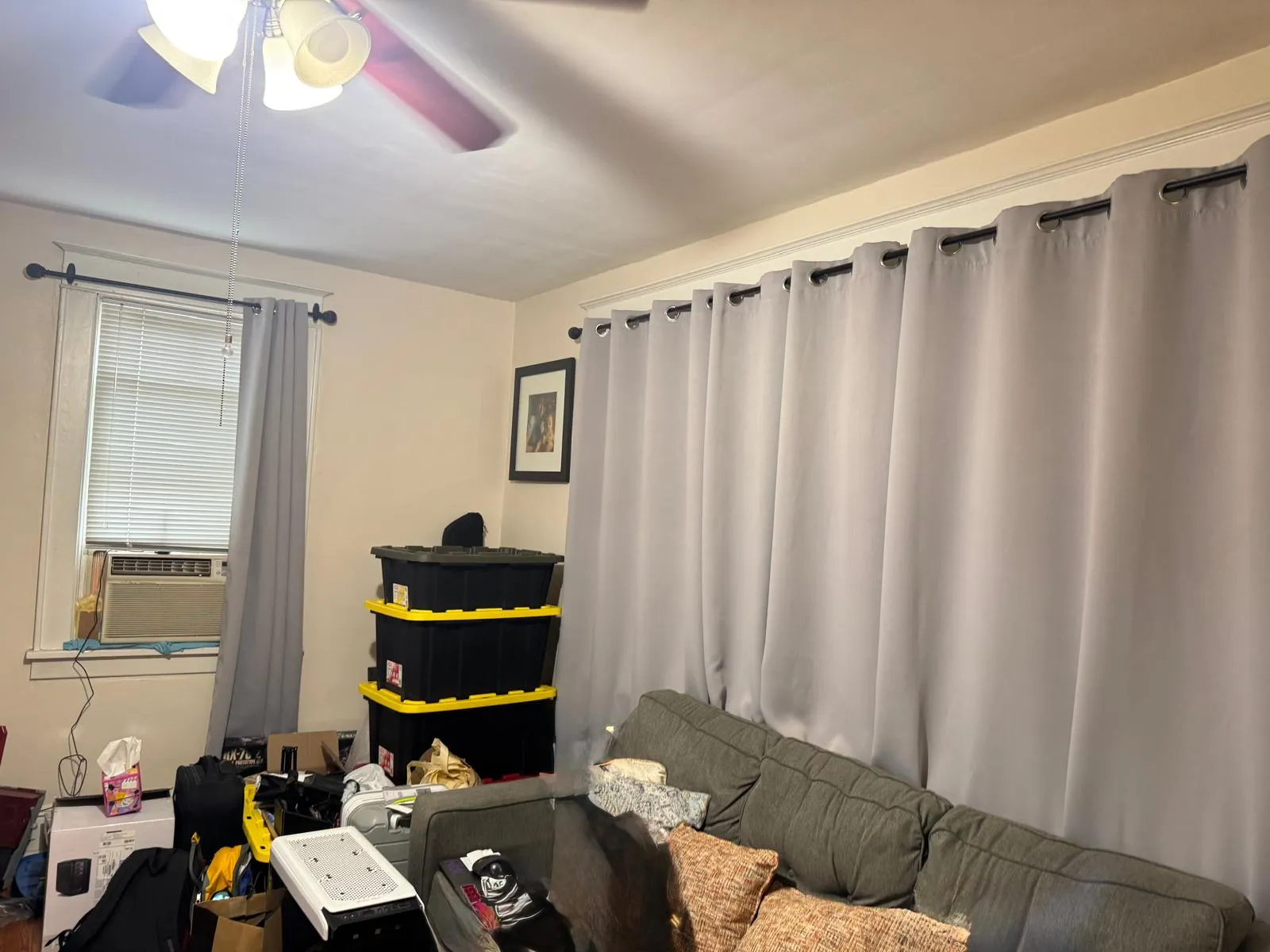 Living room before restyling: living room before: owner's couch, stacked storage bins, bags, clutter; window AC unit.