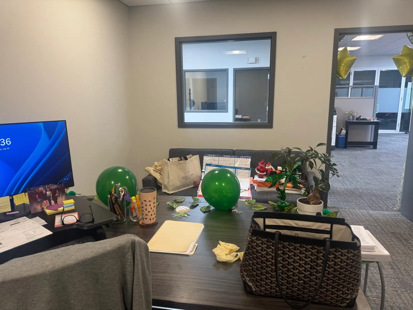 Office before restyling: executive desk with birthday balloons and designer bag; cluttered, looking toward window/sofa nook.