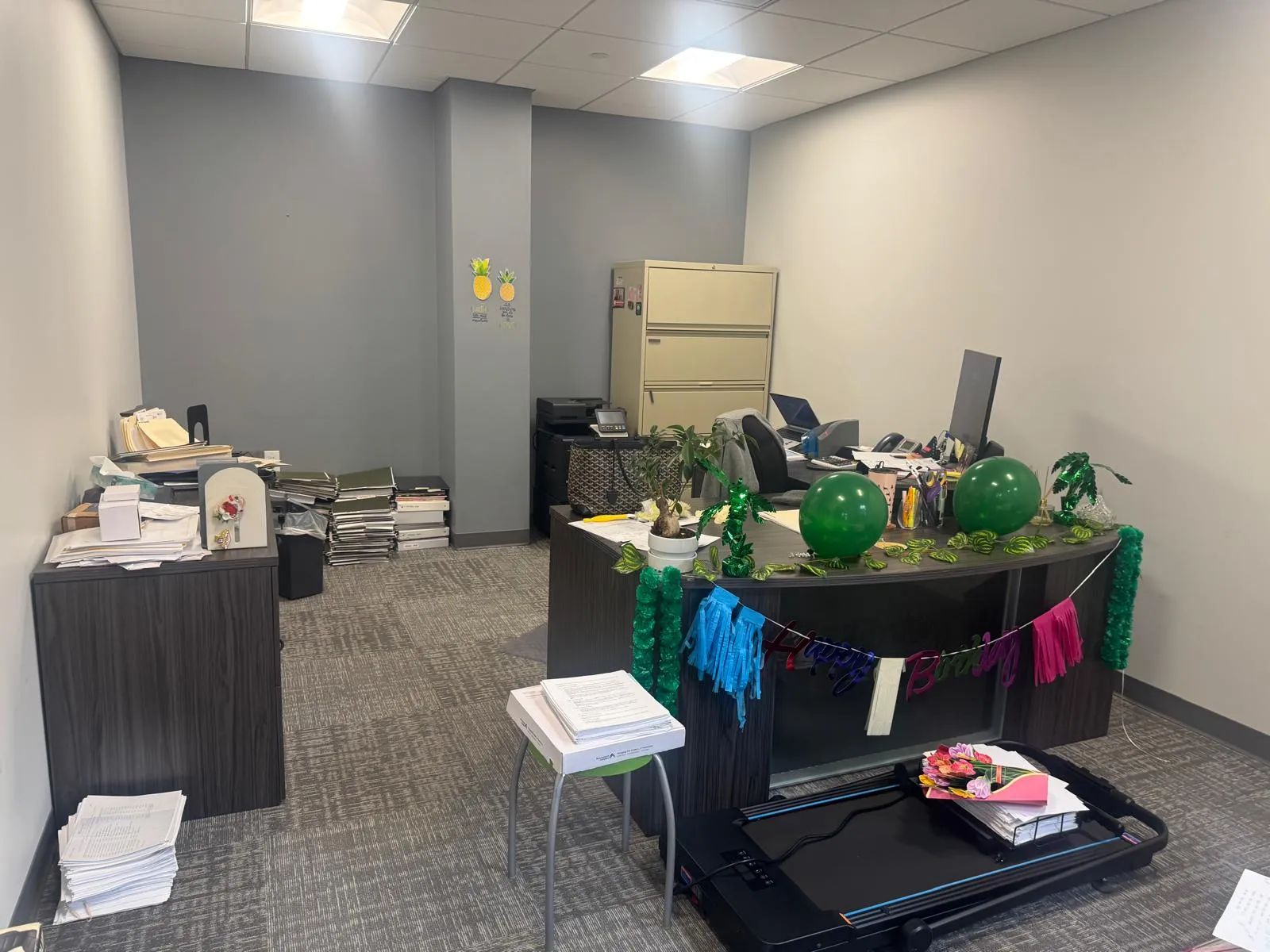 Office before restyling: wide office: 'Happy Birthday' banner, treadmill, stacked file boxes on floor.