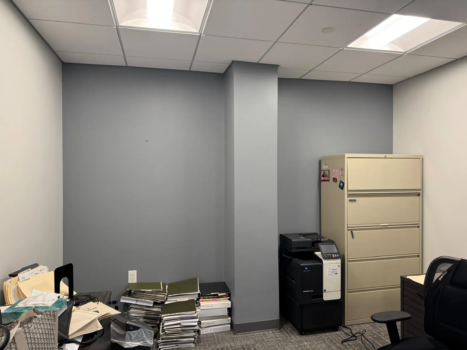 Office before restyling: corner with printer and beige lateral cabinet; binder stacks on floor, bare accent wall.