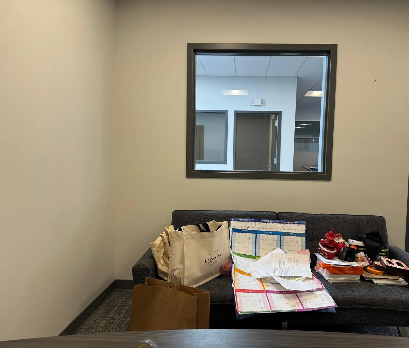 Office before restyling: sofa nook cluttered with tote/calendar/boxes; interior window behind. Cropped right side to exclude the doorway/hallway showing the client's step-and-repeat logo wall.
