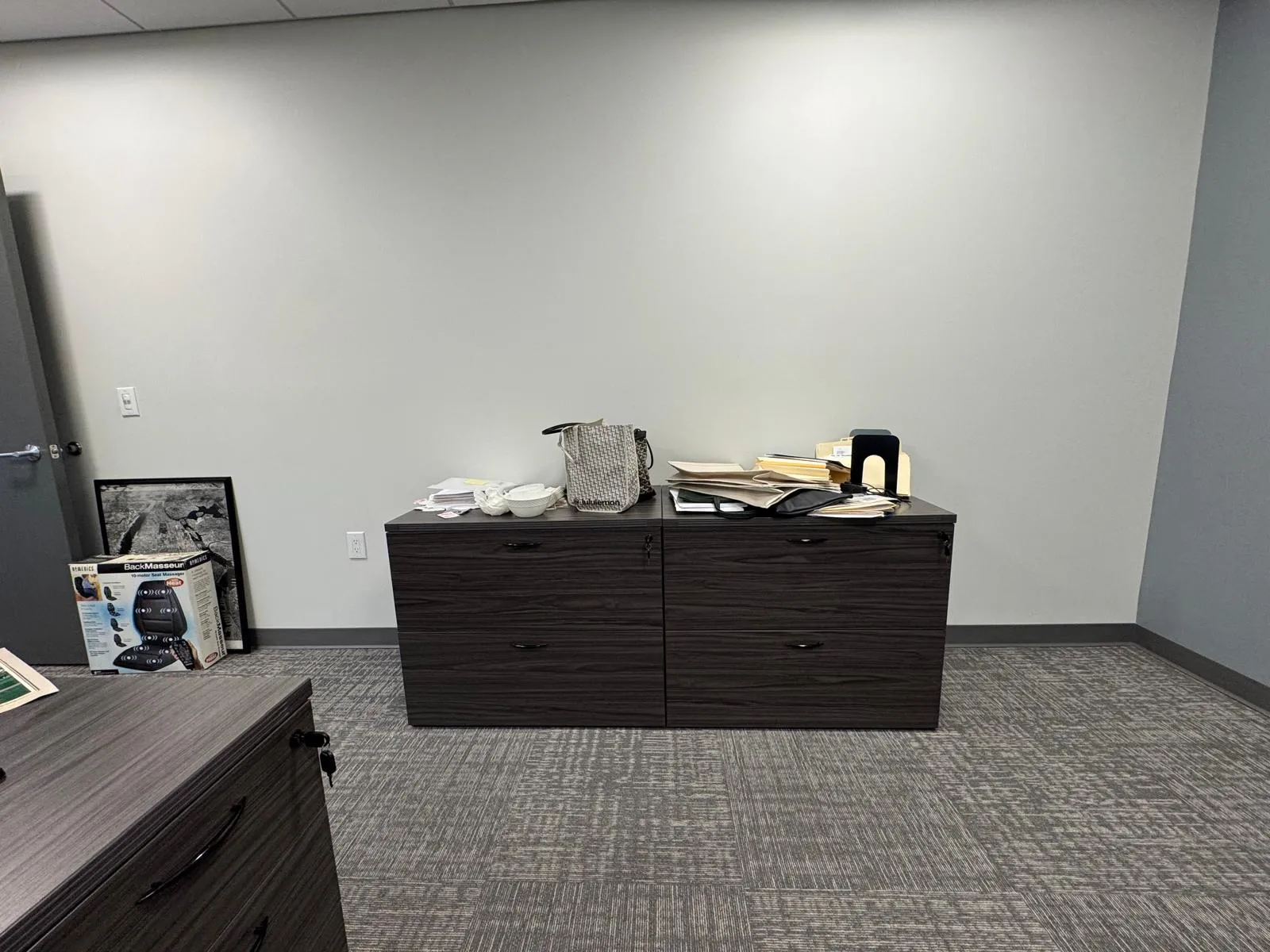 Office before restyling: credenza with massager box and shopping bags; framed motorcycle photo, bare wall.