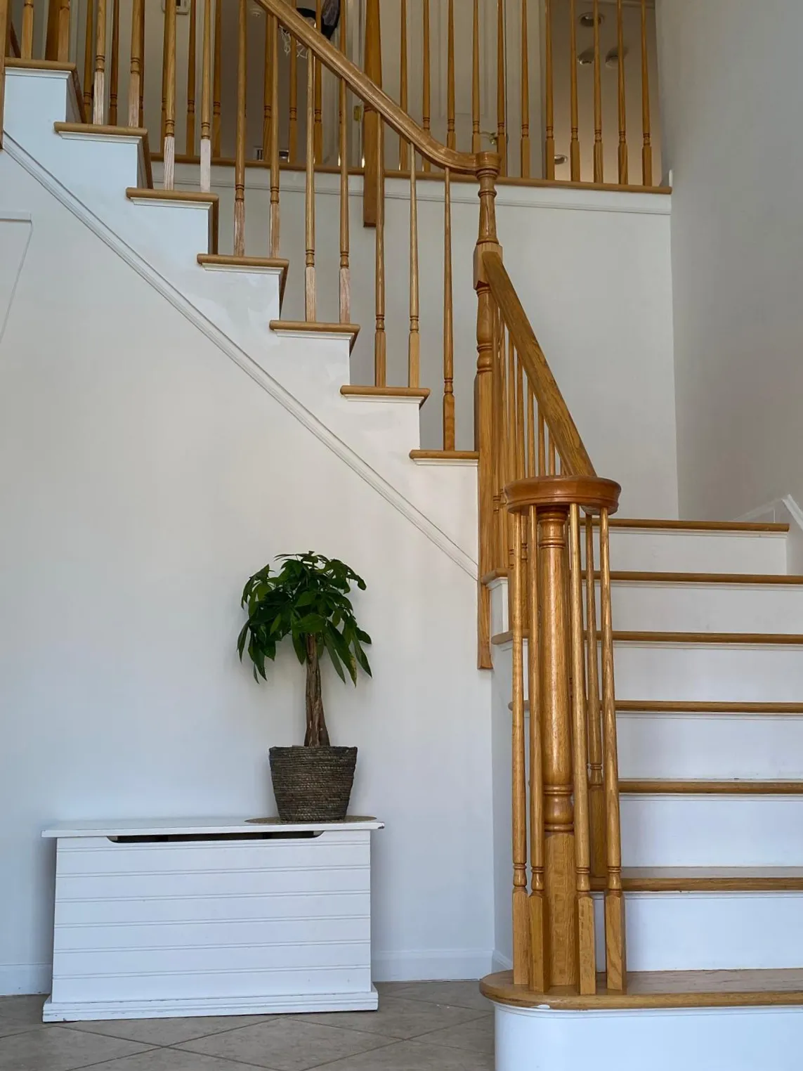 Foyer after restyling by Kultur & Decor: money tree in a woven basket on a white bench, oak balustrade rising behind.