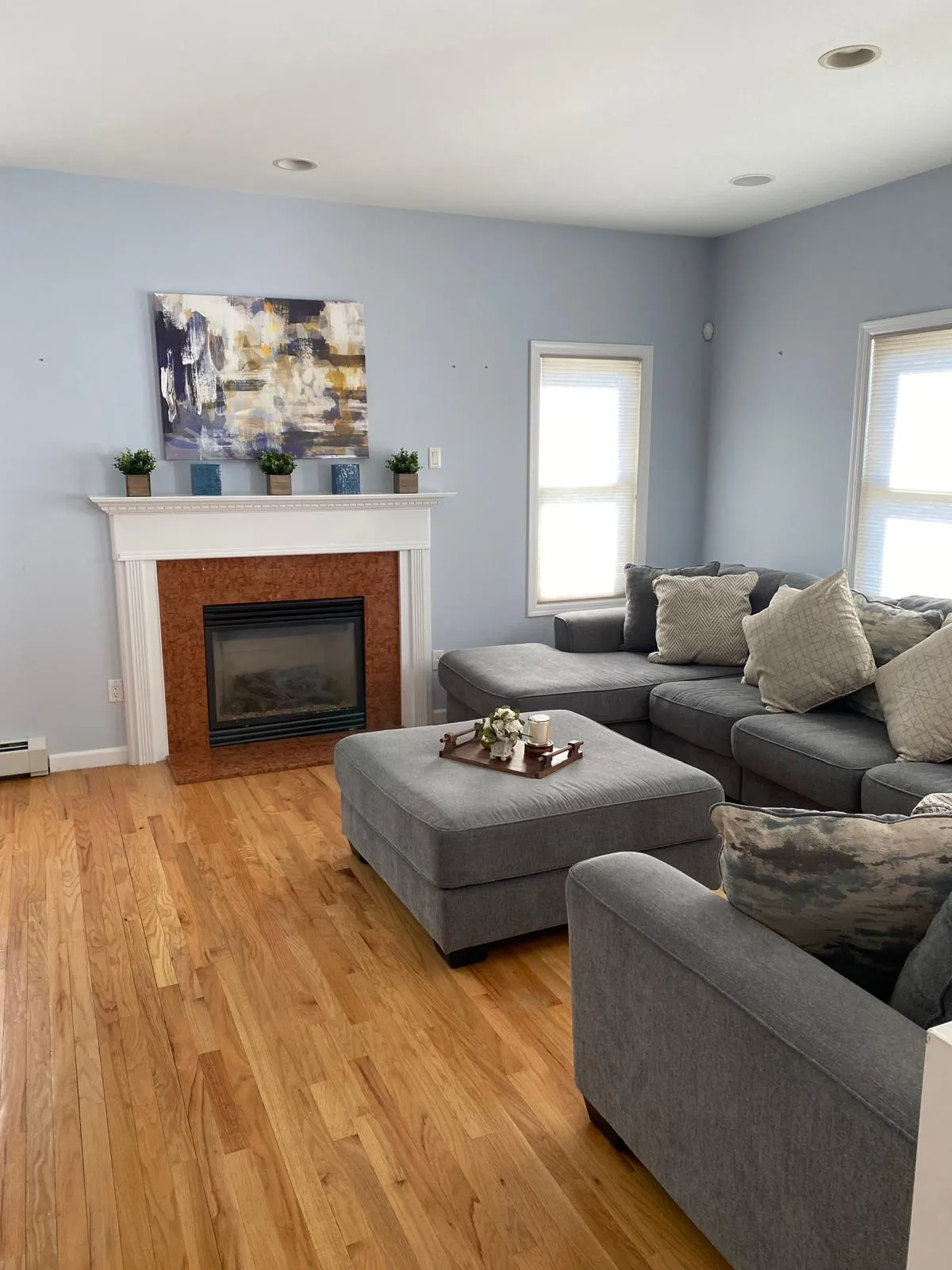 Family room after restyling by Kultur & Decor: abstract canvas over the mantel, paired greenery in wood and blue vessels, sectional and ottoman squared to the hearth.