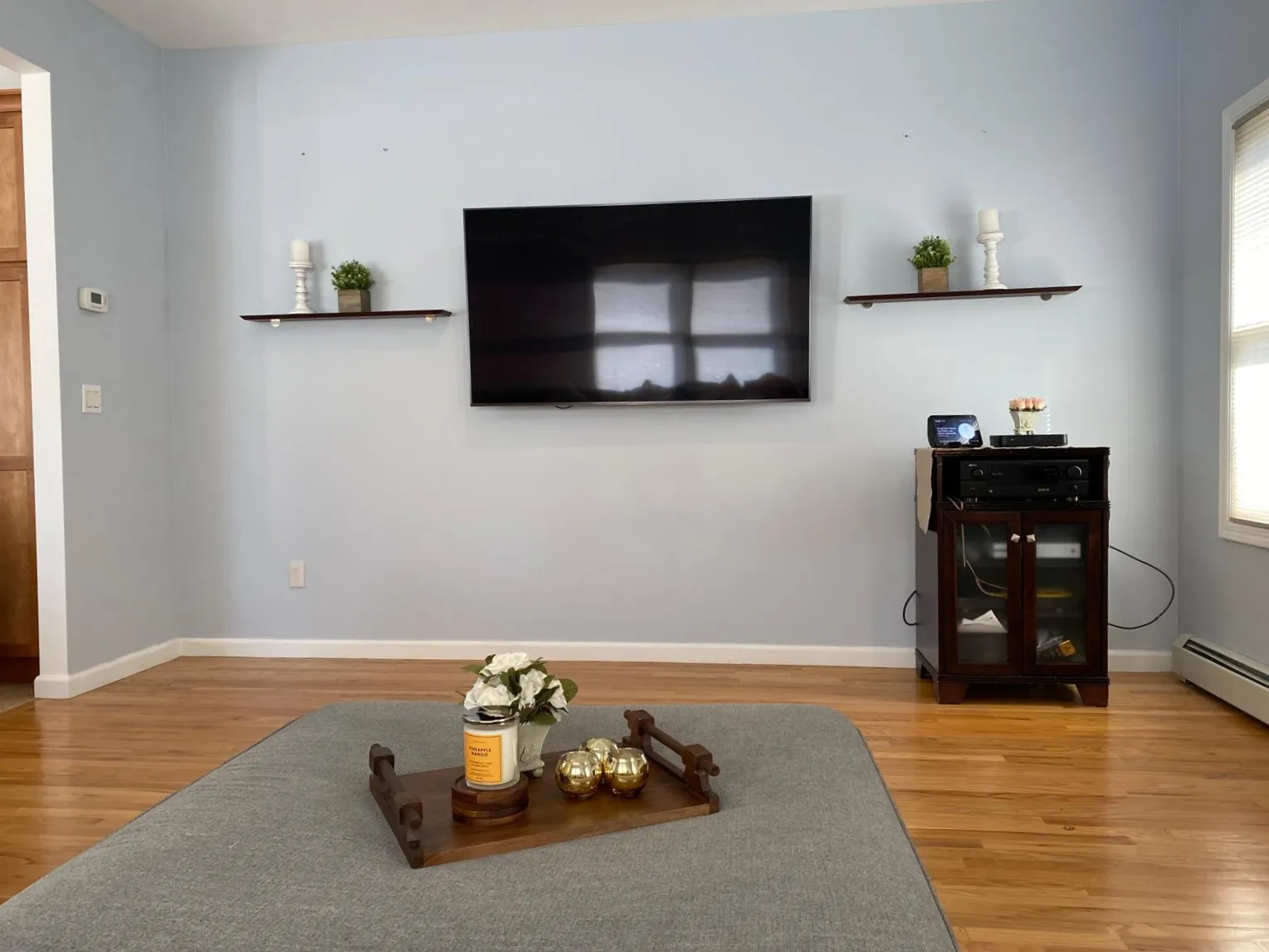 Family room after restyling by Kultur & Decor: media wall: the ottoman anchored in front of the screen and dressed with a wooden tray of gold votives and white roses, the two floating shelves either side carrying a candlestick and greenery.