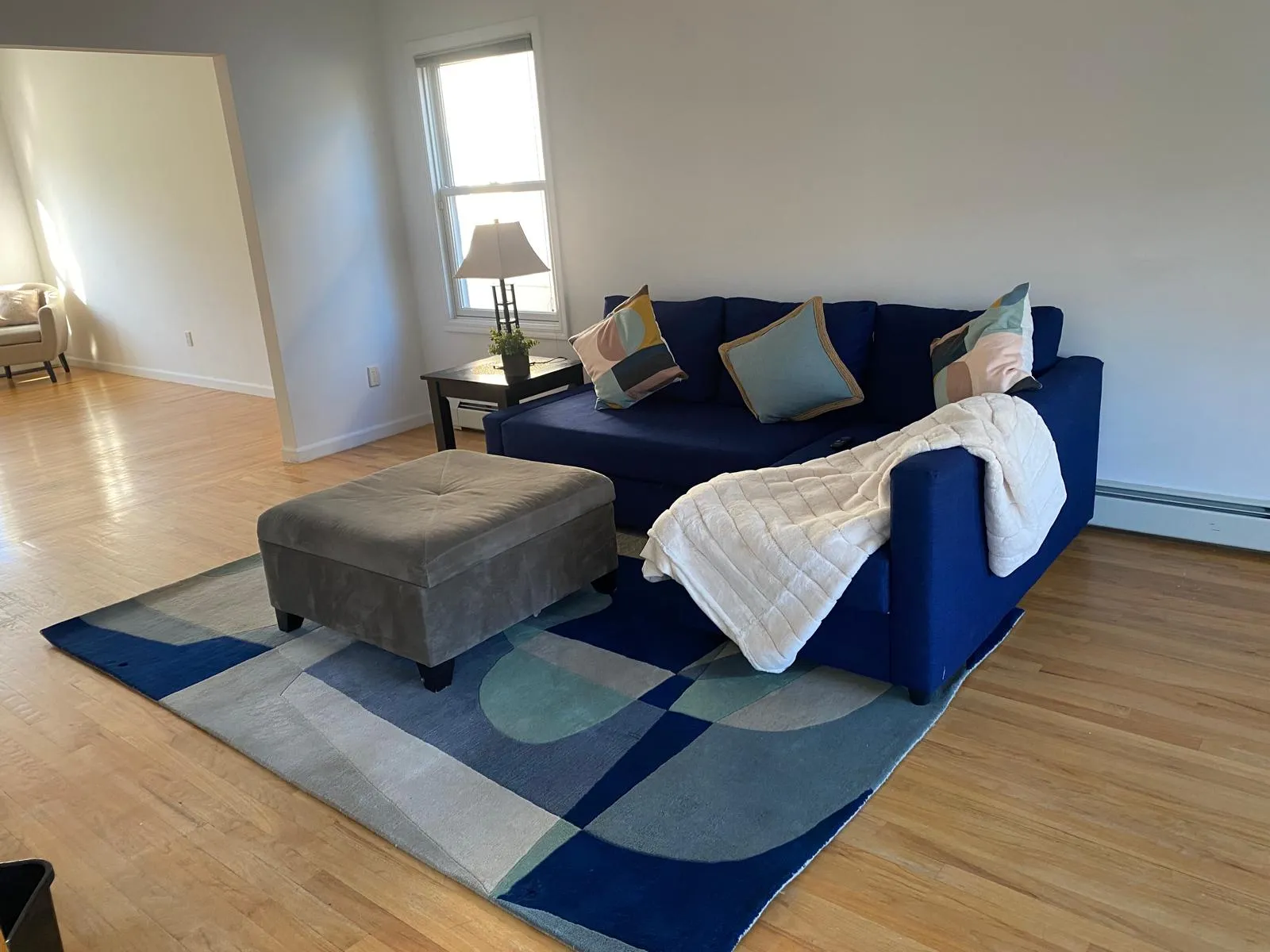 Living room after restyling by Kultur & Decor: landscape: sofa and ottoman on the rug, throw draped, the casual sitting area visible through the opening.